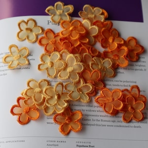 May include: A pile of orange and yellow fabric flower embellishments. The flowers are made of a sheer, delicate material and have a slightly ruffled edge.