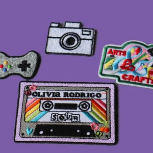May include: Four embroidered patches on a purple background. Patches include a grey video game controller, a white camera, a colourful "Arts & Crafts" design, and a cassette tape with the text "OLIVIA RODRIGO" and "SOUR".