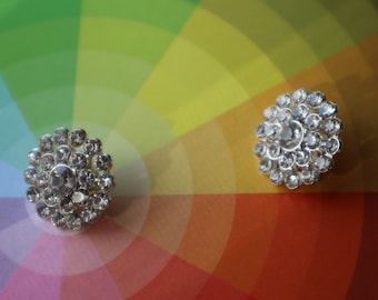 May include: Two silver-toned, round buttons with a floral design. Each button is encrusted with multiple clear, sparkling rhinestones. The buttons are set against a colorful background of a rainbow color wheel.