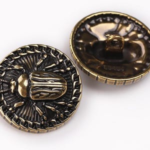 May include: Two gold-toned metal buttons with a detailed design. One button features a beetle with intricate wings and a textured background. The other button has a smooth surface.