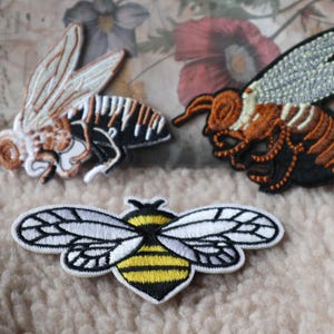 May include: Three embroidered iron-on patches featuring different bee designs. The first patch is a brown and white bee with black stripes. The second patch is a brown and orange bee with black stripes. The third patch is a yellow and black bee with white wings.