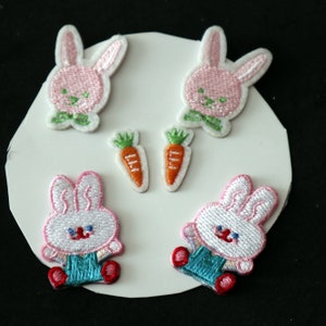 Peut inclure: Cinq patchs thermocollants représentant des lapins roses et blancs et des carottes orange. Les lapins sont brodés et les carottes sont de simples formes orange.