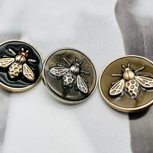 May include: Three metal buttons with a bee design. The buttons are black, silver, and gold. The bees are detailed with black, silver, and gold accents.