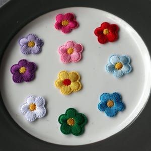 May include: A collection of ten small, five-petaled flower patches featuring intricate embroidery. The set includes vibrant colours such as magenta, red, light pink, lavender, purple, yellow with a red centre, light blue, white, dark blue, and green.
