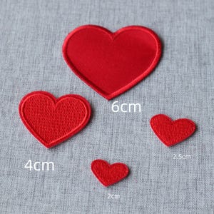 May include: Four red heart-shaped fabric patches of varying sizes. The largest heart is labelled 6cm, the medium heart is labelled 4cm, the small heart is labelled 2.5cm, and the smallest heart is labelled 2cm. Each patch has a stitched border.