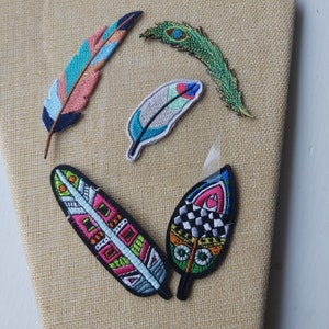 May include: Five colorful embroidered iron-on patches in the shape of feathers. The patches feature various patterns and colors, including green, blue, pink, black, and white.