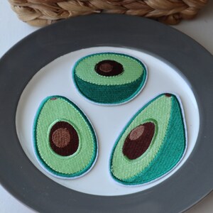 May include: Three embroidered avocado patches on a grey plate. The patches are green with brown pits and white stitching.