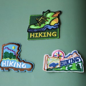 May include: Three embroidered hiking-themed patches. One shows a green hiking boot with the word "HIKING". Another depicts a blue boot with mountains and the word "HIKING". The third patch features a compass and the word "HIKING".