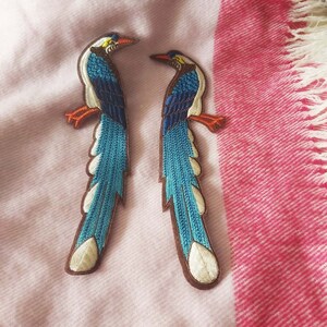 May include: Two embroidered bird patches featuring long, flowing tails in shades of bright blue, white, and brown. Each bird has a pointed orange beak, orange feet, and detailed stitching that defines the wings and plumage against a brown border.