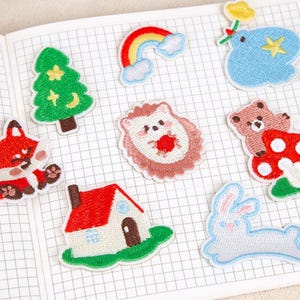 May include: A selection of embroidered patches with diverse designs. Includes a red fox, a green Christmas tree, a rainbow, a blue cloud with a star, a hedgehog, a bear on a mushroom, a house, and a white rabbit. The patches are on grid paper.