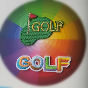 Könnte beinhalten: Zwei gestickte Aufnäher mit dem Wort "GOLF" in verschiedenen Stilen. Der obere Aufnäher ist grün mit einer roten Flagge und dem Wort "GOLF" in gelbem Garn. Der untere Aufnäher ist weiß mit dem Wort "GOLF" in Regenbogenfarben.