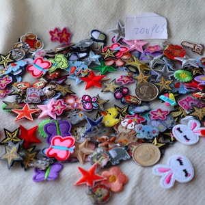 Peut inclure: Un assortiment de patchs en tissu colorés de différentes formes, notamment des étoiles, des papillons et des fleurs. Les patchs sont dans des tons de rouge, rose, jaune et bleu, certains avec des détails brodés. Un petit papier indique "200 PCS".