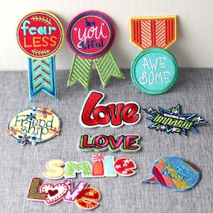 Könnte beinhalten: Sortiment an bunten gestickten Bügelbildern mit verschiedenen positiven Botschaften, darunter "Fear Less", "Be Youtiful", "Awesome", "Friendship", "Love", "Smile", "Making an Impact" und "You are the Best!"