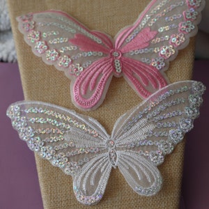 May include: Two embroidered butterfly appliques, one pink and one white, with iridescent sequins. The butterflies are on a burlap background.