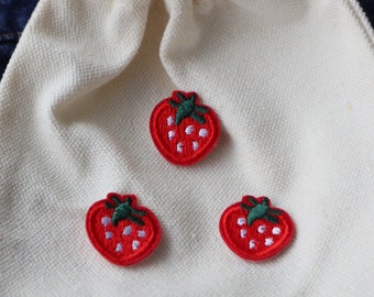 May include: Three red embroidered strawberry patches on a white fabric background.