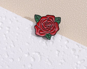 Könnte beinhalten: Rote Emaille-Rosennadel mit grünen Blättern. Die Rose hat ein detailliertes Design mit schwarzem Umriss.