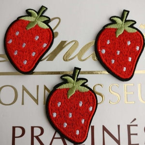 May include: Three red embroidered strawberry patches with green leaves and white seeds. The patches are on a white background with brown text that reads "PRALINÉS".