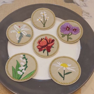 May include: Six embroidered flower patches on a beige background. The patches are circular and have a gold trim. The flowers include a white lily, a red peony, a white daisy, a purple flower, and two white flowers with yellow centres.