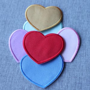 May include: A collection of six heart-shaped fabric patches arranged in a cluster. The patches feature solid colours including red, light blue, tan, lavender, and light purple, each finished with a matching stitched border around the edges.