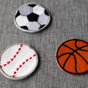 Peut inclure: Trois patchs brodés représentant un ballon de baseball, un ballon de basket-ball et un ballon de football. Le ballon de baseball est blanc avec des coutures rouges, le ballon de basket-ball est orange avec des coutures noires et le ballon de football est noir et blanc.