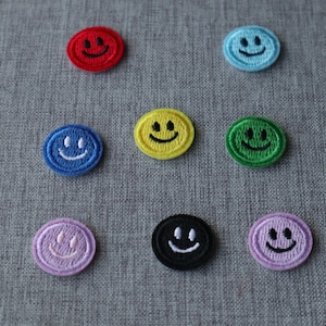 May include: A selection of embroidered smiley face patches in a range of colours, such as red, blue, yellow, green, black, and purple. Each patch has a smiling face with a black stitched outline. The patches are round and approximately 2.5 cm in diameter.