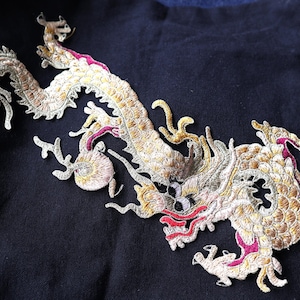 Könnte beinhalten: Gold gesticktes Drachenmotiv auf schwarzem Stoff. Der Drache ist mit goldenen und roten Fäden verziert.