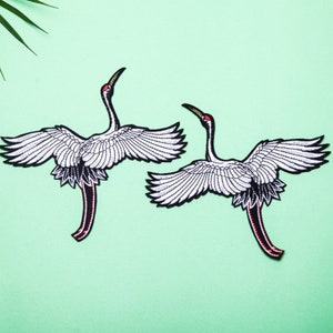 May include: Two white embroidered iron-on patches of cranes with black and red accents. The cranes are in flight with their wings spread.