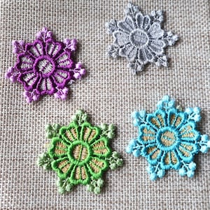 May include: Four decorative lace flowers in different colors: purple, gray, green, and blue. The flowers are on a burlap background.