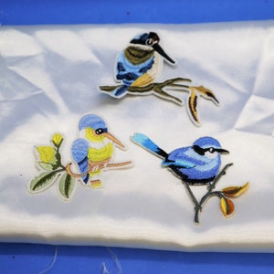 May include: Three embroidered iron-on patches featuring blue birds with yellow and white accents perched on branches with green leaves.