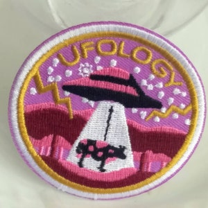 May include: Embroidered patch with a purple background and the word "UFOLOG" in gold lettering. The patch features a black and pink UFO beaming a cow with pink spots. Lightning bolts and pink hills complete the design.