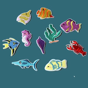 Peut inclure: Un ensemble de onze écussons en tissu brodé représentant divers animaux marins et éléments océaniques, incluant un homard rouge, un hippocampe violet, une algue verte, un coquillage violet et plusieurs motifs de poissons tropicaux colorés.