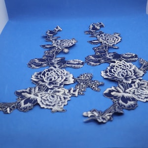 May include: Two white floral lace appliques with a navy blue outline. The appliques are on a blue background.