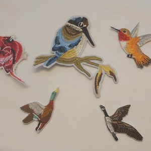 May include: Embroidered patches featuring various birds: a red cardinal, a blue and brown bird perched on a branch, a hummingbird, a duck, and a goose. Each patch has detailed stitching and a white border.