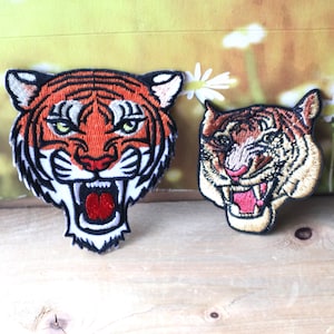 May include: Two embroidered tiger face patches. The larger patch is orange and black with a white background. The smaller patch is brown and black with a white background. Both patches have a fierce expression with open mouths and sharp teeth.