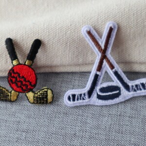 Peut inclure: Deux patchs brodés à repasser. L'un représente un ballon de cricket rouge et blanc avec deux chauves-souris croisées. L'autre représente deux bâtons de hockey croisés avec une rondelle.
