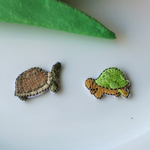May include: Two embroidered iron-on patches of turtles. One turtle is brown with a green shell and the other is green with a brown shell.