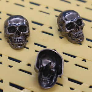 May include: Three silver skull-shaped buttons. The skulls have detailed features, including eye sockets, a nose, and a mouth with teeth.