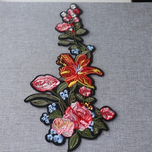 Peut inclure: Un écusson floral brodé présentant un grand lys orange au centre, des roses roses et rouges, ainsi que de petites fleurs bleu clair. Le motif comprend des feuilles et des tiges vert foncé disposées en grappe verticale sur un fond noir.