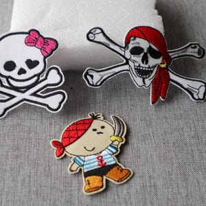 May include: Three embroidered patches with pirate themes. One patch features a skull with a pink bow and crossbones. Another shows a skull with a red bandana and crossbones. The third patch depicts a cartoon pirate with a sword.