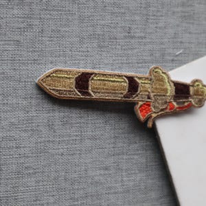 May include: An embroidered patch shaped like a sword with a gold blade, dark brown accents, and a hilt featuring a bright orange detail. The design is rendered in dense thread work with a defined border, showcasing a metallic gold sheen.
