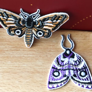 May include: Two embroidered iron-on patches featuring moth designs. One moth is brown and black with white eyes. The other moth is purple and white with black accents.
