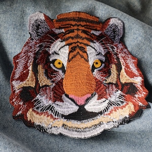 May include: Embroidered tiger patch with a detailed design featuring orange, brown, white, and black colours. The tiger's face has yellow eyes and a pink nose. The patch is placed on a light blue denim fabric.