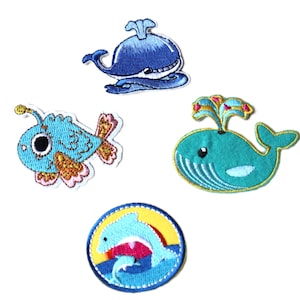 May include: Set of four embroidered iron-on patches featuring ocean creatures. The patches include a blue whale, a turquoise whale, a blue fish with orange and pink accents, and a blue dolphin jumping over a wave.