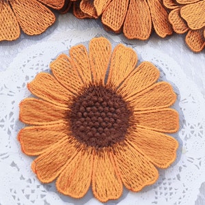 Peut inclure: Applique brodée de tournesol orange avec un centre brun. Le tournesol est sur un fond de dentelle blanche.