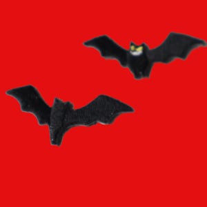 Peut inclure: Deux petits ornements ou appliques en forme de chauve-souris noire. La chauve-souris du haut présente des yeux jaunes visibles et un détail de bouche blanche, tandis que celle du bas est entièrement noire avec des ailes texturées. Les deux sont en vol.