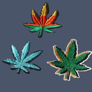 Peut inclure: Trois écussons brodés en forme de feuilles de cannabis. L'écusson du haut présente un dégradé de jaune, d'orange et de vert. L'écusson en bas à gauche est bleu clair uni. L'écusson en bas à droite est vert avec des reflets jaunes et une bordure blanche.