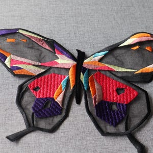 May include: Embroidered butterfly patch with a black mesh. The butterfly has colourful geometric patterns in red, orange, purple, pink, and blue. The patch is designed for decorative purposes, ideal for clothing or accessories.