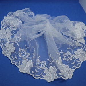 May include: White tulle fabric with a delicate floral lace trim. The lace features a repeating pattern of small white flowers and leaves.
