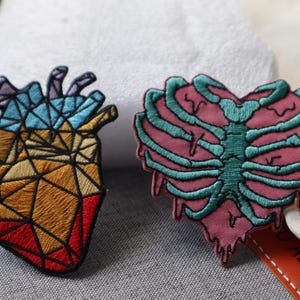 May include: Two embroidered patches: a geometric heart in red, orange, blue, and purple, and a rib cage in pink and teal. These patches are likely for clothing or accessories.