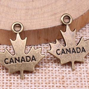 May include: Two bronze-colored maple leaf charms. Each charm has the word "CANADA" inscribed and a small loop at the top for attaching to jewelry or other crafts. The charms are on a wooden surface.